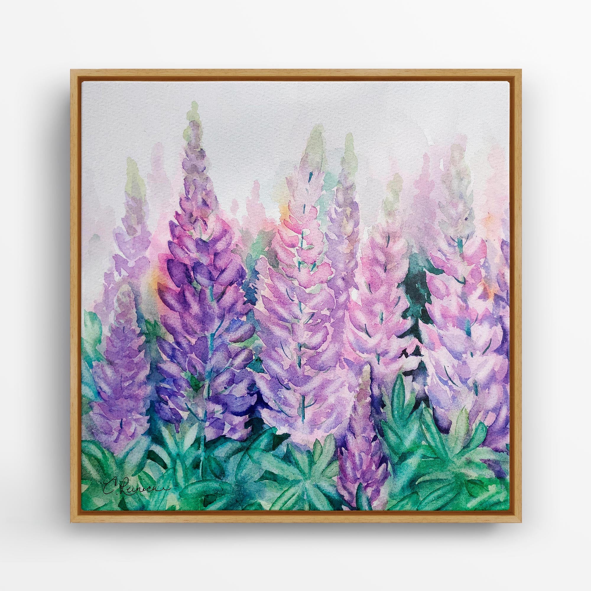 A wood-framed wall art canvas of a lupine wildflower print. The print is of a group of lupine wildflowers in various shades of purple and blue. The flowers are in a field of green leaves. The frame is a light wood color, and the background is white. The image is signed by the artist, Conni Reinecke in the bottom left corner.